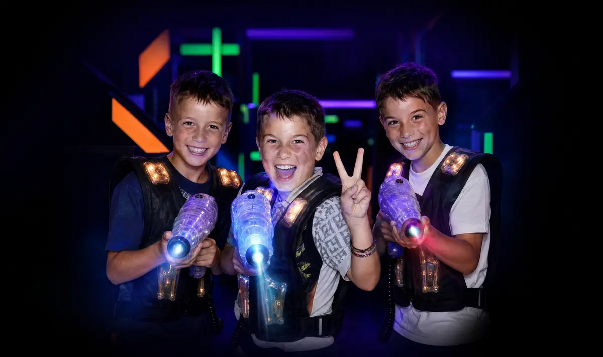 Three smiling kids with glowing vests and blasters in the black-light maze